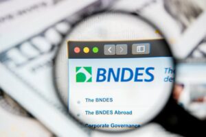 Imagem de uma lupa focada na tela de um computador com o logo do BNDES destacando-se na pesquisa online.
