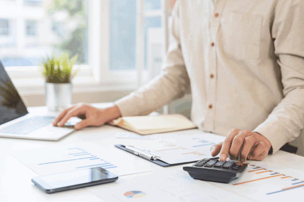 Profissional utilizando calculadora e computador para analisar planilhas e gráficos de custos em um ambiente de escritório