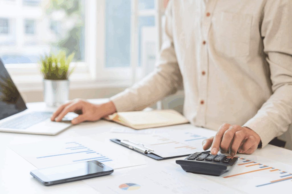 Profissional utilizando calculadora e computador para analisar planilhas e gráficos de custos em um ambiente de escritório