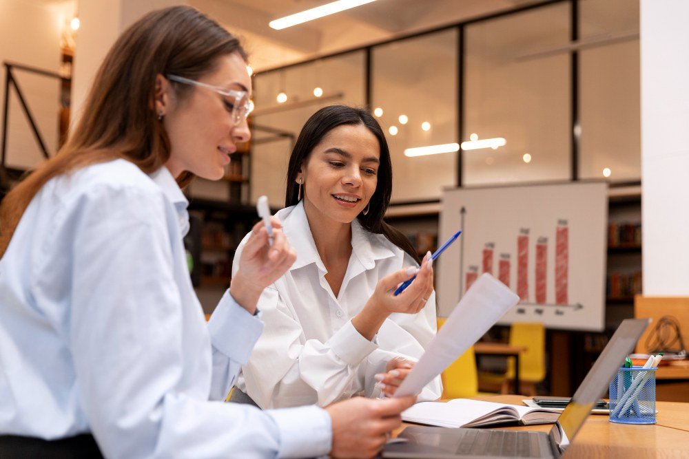 Duas mulheres profissionais discutindo projetos e estratégias de negócios em uma reunião de trabalho, com gráficos e laptops na mesa.