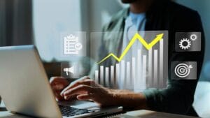 Pessoa digitando no notebook enquanto um gráfico ascendente relacionado à análise de custos aparece em destaque.