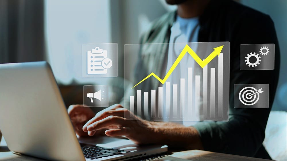 Pessoa digitando no notebook enquanto um gráfico ascendente relacionado à análise de custos aparece em destaque.